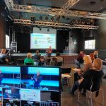 Kuvassa sali, jossa etualalla on kaksi televisioruutua, joissa näkyy luennoitsija. Kuvaruutujen taustalla on ryhmä opiskelijoita, jotka istuvat salissa maskit kasvoillaan. Sama luennoitsija, joka näkyy kuvaruuduissa, puhuu salissa häntä kuunteleville opiskelijoille. 