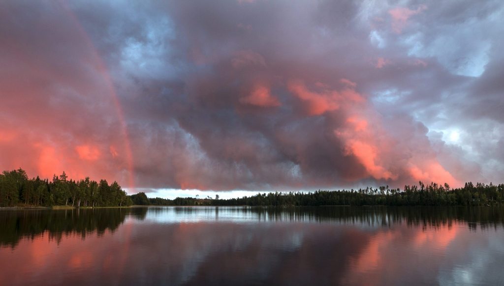 Kuvituskuva.Myrskysään tummentamat pilvet roikkuvat valokuvassa auringon punertamina peilityynen järven yllä. Järven rantoja kiertää metsä. Horisontissa metsän ja pilvien välistä kajastaa kesäöinen valo. Pilvien välissä on aavistus korkeuksiin ulottuvasta sateenkaaresta.